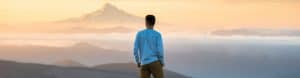 Pilgrim stands on a mountain at dusk with another mountain summit in the distance.