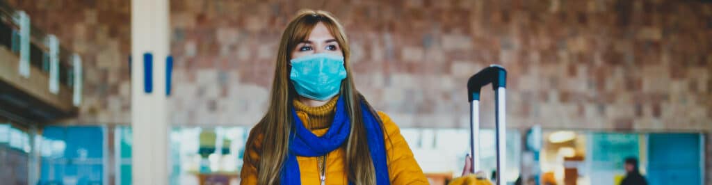 Traveling During COVID - A woman wearing a mask waits at a station with her luggage.